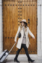 Glückliche Frau mit Mantel, die ihr Gepäck im Gehen an der Tür entlang rollt - EBBF02078