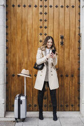 Smiling mid adult woman with wheeled luggage using mobile phone against wood door - EBBF02079