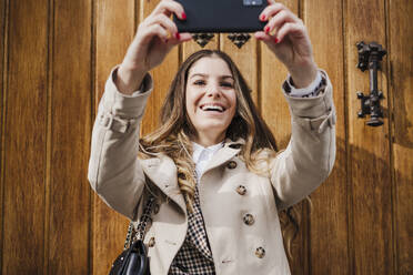 Cheerful woman taking selfie while standing against wood door - EBBF02080