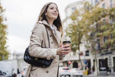Lächelnde Frau, die eine Einweg-Kaffeetasse hält, während sie auf einer Straße in der Stadt wegschaut - EBBF02087
