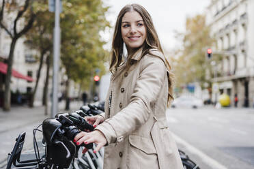 Modische Frau schaut weg, während sie ein Elektrofahrrad an einer Parkstation hält - EBBF02088