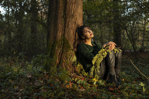 Schöne Frau mit Zweig träumt Tag, während lehnt sich auf Baumstamm in öffentlichen Park - AXHF00029
