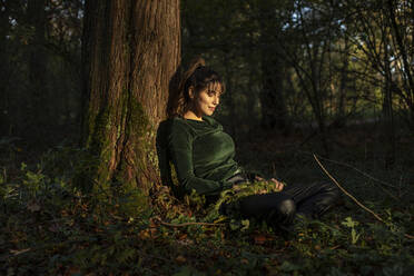 Mittlere erwachsene Frau lehnt sich an einen Baumstamm im Sonnenlicht des Herbstes - AXHF00030