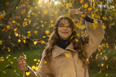 Mid adult woman with eyes closed holding branch in public park during autumn - AXHF00040