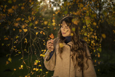 Beautiful woman wearing warm jacket looking at autumn leaf in park - AXHF00042