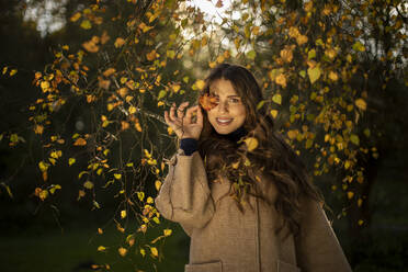 Lächelnde Frau, die ihr Auge vor einem Herbstblatt im öffentlichen Park verdeckt - AXHF00043