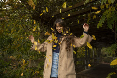 Glückliche Frau mit erhobenen Armen, die in einem öffentlichen Park im Herbst in fallenden Blättern steht - AXHF00045