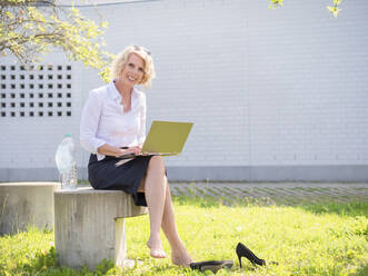 Smiling businesswoman using laptop while sitting on seat against wall - LAF02626