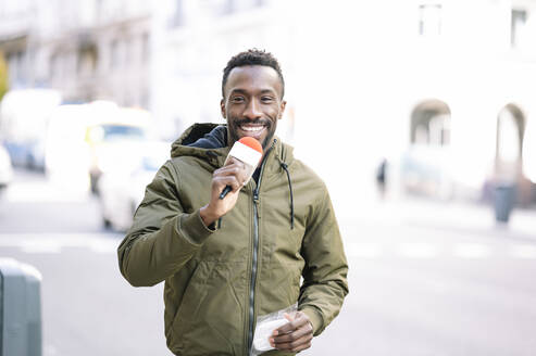 Lächelnder männlicher Journalist mit Mikrofon auf der Straße in der Stadt - JCMF01770