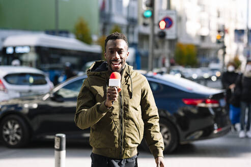 Männlicher Fernsehreporter spricht über Mikrofon, während er auf der Straße steht - JCMF01776