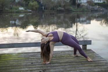 Junge Frau beugt sich nach hinten und übt Yoga auf dem Pier gegen den See bei Sonnenuntergang - AXHF00057