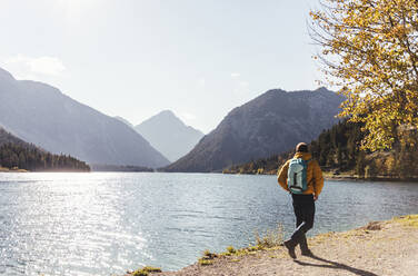 Male hiker with backpack walking by lake against sky during sunny day - UUF22429