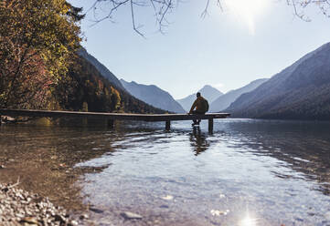 Silhouette male hiker sitting on pier over lake against sky during sunny day - UUF22431