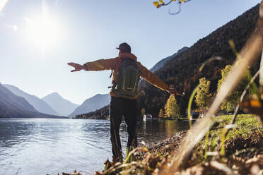 Mature male hiker with arms outstretched looking at lake against clear sky during sunny day - UUF22433