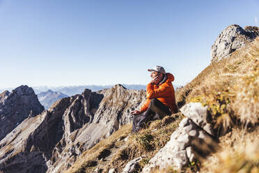 Männlicher Wanderer, der auf einem Berg sitzend gegen den klaren Himmel an einem sonnigen Tag nachdenkt - UUF22438