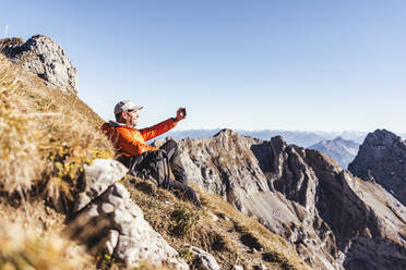 Mature man taking selfie with smart phone while sitting on mountain against clear sky - UUF22440