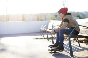 Junger Junge mit Kapuze sitzt auf einer Bank mit Schuhen über dem Skateboard - CAVF91566