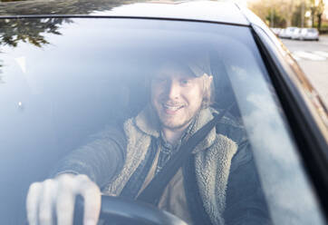 Lächelnder erwachsener Mann am Steuer eines Autos, gesehen durch die Windschutzscheibe - JCCMF00628