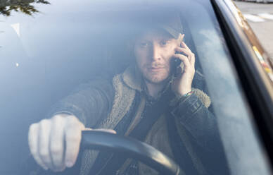 Mittlerer erwachsener Mann, der über ein Smartphone spricht, während er Auto fährt, gesehen durch die Windschutzscheibe - JCCMF00630