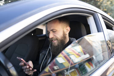 Bearded man using mobile phone while sitting in car seen through window - JCCMF00637