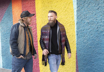 Gay boyfriends talking while walking against colorful wall in city - JCCMF00639