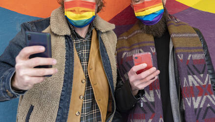 Schwule Freunde mit Regenbogenmasken benutzen Smartphones, während sie an der Wand stehen - JCCMF00644