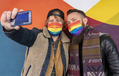 Schwule Freunde mit Regenbogenmasken machen ein Selfie mit ihrem Smartphone an der Wand - JCCMF00645