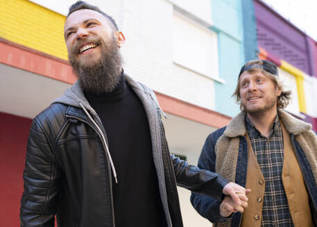 Lächelnde Homosexuell Paar hält Hände beim Gehen gegen bunte Gebäude in der Stadt - JCCMF00648