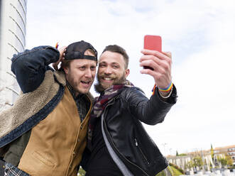 Fröhlicher Mann, der ein Selfie mit seinem Freund macht, während er gegen den Himmel steht - JCCMF00668