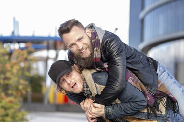 Cheerful gay man piggybacking boyfriend while standing in city - JCCMF00676