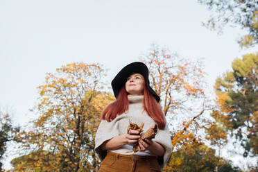 Thoughtful woman wearing hat holding autumn leaves while standing against clear sky in park - MRRF00775