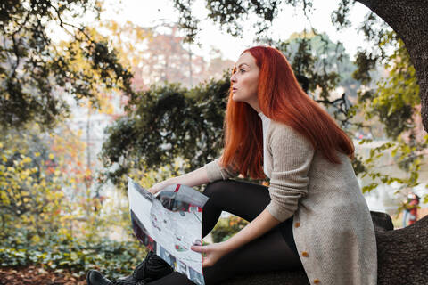 Schöne Frau mit braunen Haaren, die eine Karte hält, während sie im Park sitzt, lizenzfreies Stockfoto