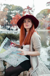 Beautiful woman wearing hat holding map while sitting in park - MRRF00779