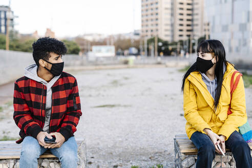 Couple wearing protective face mask sitting at social distance on bench during COVID-19 - JCZF00400