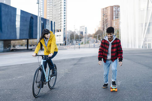 Lächelnde Frau auf dem Fahrrad neben einem Freund, der auf der Straße Skateboard fährt - JCZF00405
