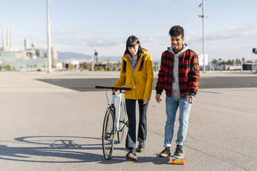 Junge Frau mit Fahrrad geht an einem Freund vorbei, der auf der Straße Skateboard fährt - JCZF00406