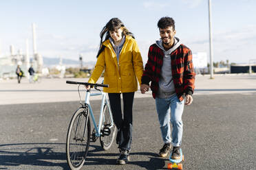 Lächelnder Mann fährt Skateboard und hält die Hand eines Freundes, der mit dem Fahrrad auf der Straße fährt - JCZF00407