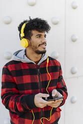Lächelnder Mann mit Kopfhörern und Mobiltelefon, der wegschaut, während er an der Wand steht - JCZF00409