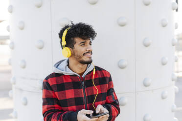 Junger Mann mit Kopfhörern, der ein Mobiltelefon benutzt, während er an der Wand steht - JCZF00410
