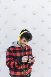 Young man smiling while using mobile phone standing against wall - JCZF00412