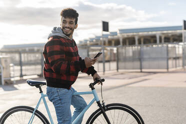 Lächelnder Mann mit Mobiltelefon, der wegschaut, während er an einem sonnigen Tag auf dem Fahrrad steht - JCZF00417
