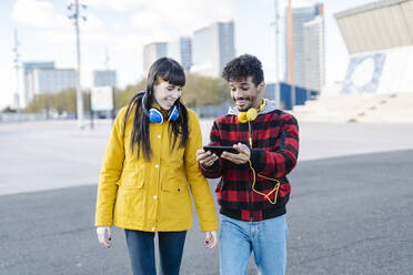 Smiling couple wearing headphones using mobile phone while walking on road - JCZF00423