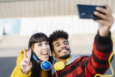 Lächelnder Mann, der ein Selfie über sein Handy mit einem Freund macht, der ein Friedenszeichen zeigt, während er im Freien steht - JCZF00424