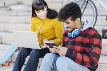 Junger Mann, der ein digitales Tablet benutzt, während er neben einer Freundin sitzt, die einen Laptop auf einer Treppe benutzt - JCZF00435
