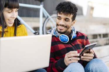 Lächelnde Freunde, die im Freien sitzend einen Laptop und ein digitales Tablet benutzen - JCZF00437