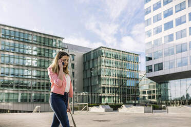 Spanien, Barcelona, Junge Geschäftsfrau vor Bürogebäude gehend, telefonierend - XLGF00946