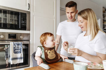 Mother feeding baby daughter at kitchen table - VYF00380