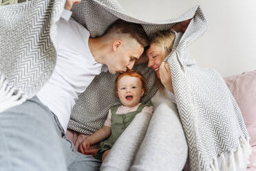 Happy family with baby daughter hiding under blanket on sofa - VYF00385