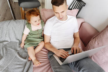 Mann mit kleiner Tochter arbeitet am Laptop auf dem Sofa - VYF00396