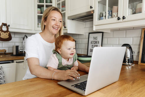 Frau mit kleiner Tochter benutzt Laptop am Küchentisch - VYF00401